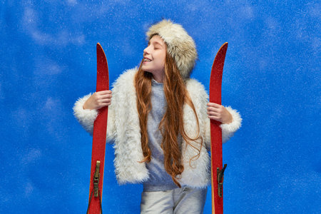 winter joy, pleased preteen girl in faux fur jacket and hat holding red skis on turquoise backgroundの写真素材