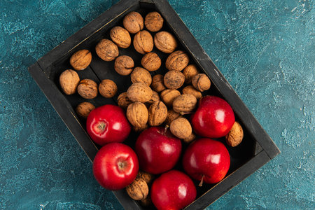 red ripe apples and whole walnuts in black wooden tray on blue textured table, thanksgiving conceptの写真素材