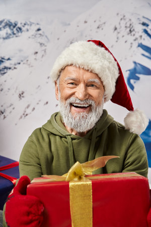portrait of bearded Santa holding big red present wearing red hat and mittens, Christmas conceptの写真素材