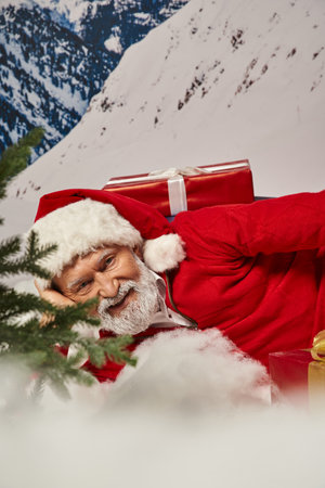 close up of joyous Santa in christmassy hat lying on snow surrounded by presents, winter conceptの写真素材