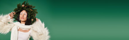 holiday banner, brunette asian woman with white makeup and trendy outfit posing in wreath on green