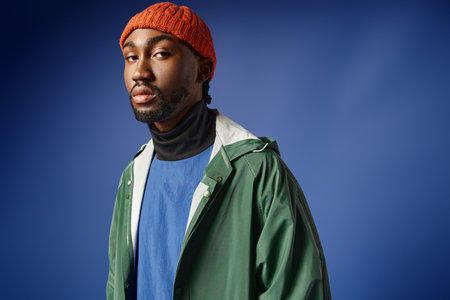 stylish african american man in knitted hat and trendy winter attire posing on blue backdrop