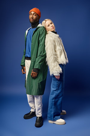 blonde woman leaning on african american man in winter attire, diverse young couple on blue backdropの写真素材
