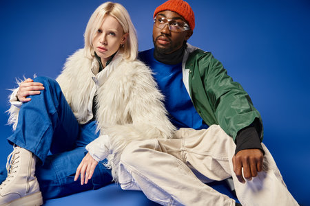 young interracial couple in winter attire sitting together on blue backdrop, diversity and fashion