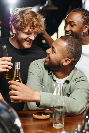 three joyful multiethnic friends smiling and toasting glasses of beer while spending time in barの写真素材