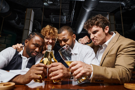 four multicultural colleagues in formal wear toasting with beer in bar, men spending time after work