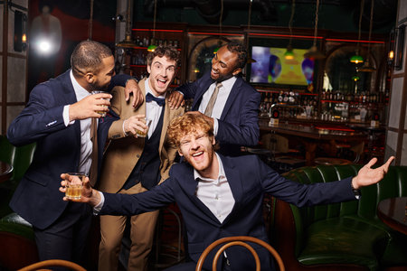 happy interracial friends in formal wear having bachelor party and holding whiskey in bar, diversityの写真素材