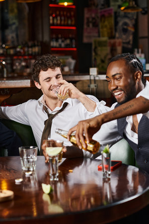 happy drunk man drinking whiskey near cheerful african american friend pouring beer in glassの写真素材