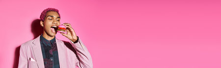 stylish handsome man with pink curly hair with earrings eating mini burger on pink backdrop, bannerの写真素材