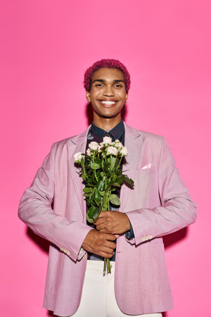 cheerful man with curly hair in pink blazer posing with rose bouquet and looking at cameraの写真素材
