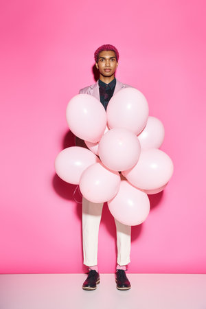 handsome man with pink hair holding pink balloons and looking at camera posing on pink backdropの写真素材