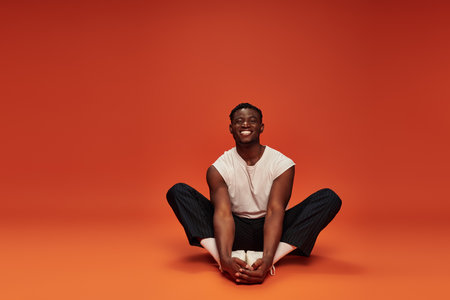 overjoyed african American man in white tank top and black pants sitting on red and orange backdropの写真素材