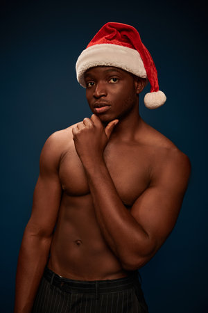 thoughtful african american man in santa hat posing with shirtless torso on dark blue backdropの写真素材