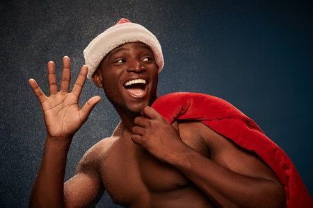 overjoyed sportive african american man with santa bag waving hand on navy blue snowy backdropの写真素材