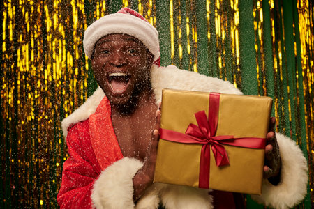 energized african american man in santa costume with christmas bag and open mouth under snowfallの写真素材
