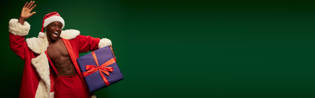 hot african american guy in santa claus costume holding present and waving hand on green, bannerの写真素材