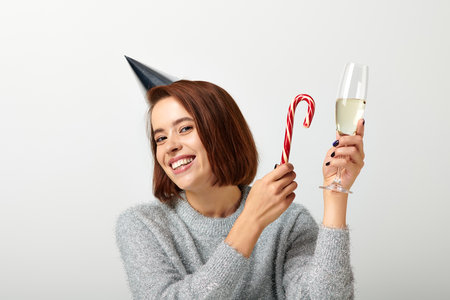 positive woman in party cap holding champagne glass and candy cane on grey, Merry Christmasの写真素材