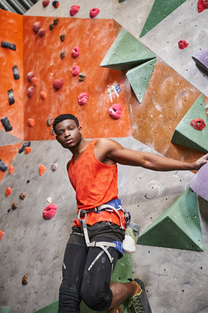 handsome african american man with alpine harness in orange shirt posing next to rock climbing wallの写真素材
