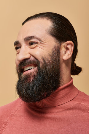 positive man with beard in pink turtleneck jumper smiling cheerfully on beige backdrop