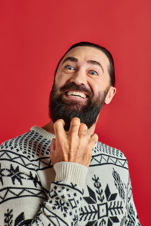 passionate bearded man in Christmas sweater with ornament smiling on red backdrop, winter holidaysの写真素材