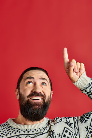 happy bearded man in Christmas sweater with ornament pointing up on red backdrop, winter holidaysの写真素材