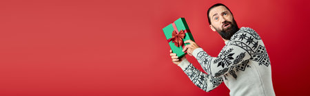 confused bearded man in winter sweater with ornament holding Christmas present on red, banner