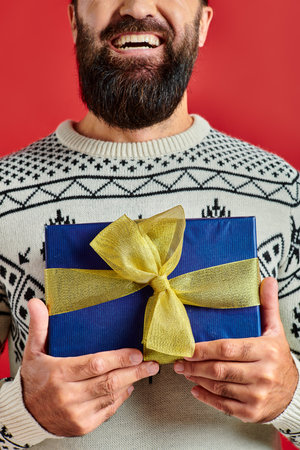 cropped happy bearded man in winter sweater with ornament holding Christmas present on red backdropの写真素材