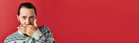 bearded man in Christmas sweater with ornament covering mouth on red, winter holidays bannerの写真素材