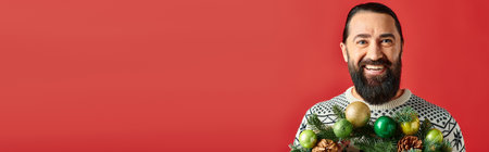 happy bearded man in winter sweater holding decorated Christmas wreath with baubles on red, bannerの写真素材