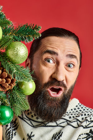 positive bearded man in winter sweater holding Christmas wreath with baubles on red backdropの写真素材