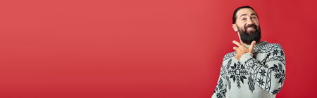 happy bearded man in Christmas sweater with ornament smiling on red backdrop, winter holidaysの写真素材