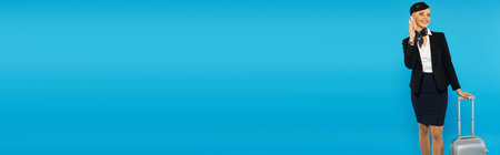 elegant air hostess in uniform walking with travel bag and talking on smartphone on blue, bannerの写真素材