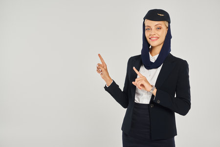 charming stewardess in arabian airlines uniform smiling at camera and pointing with fingers on greyの写真素材