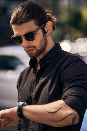 vertical shot of good looking male model in elegant black outfit looking at his wristwatch, fashionの写真素材