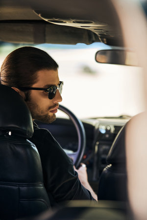 vertical shot of good looking male model in black elegant attire at steering wheel, turning his head