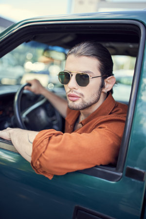 appealing young man with earring in trendy brown shirt with sunglasses looking out of car windowの写真素材