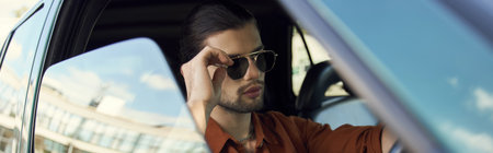 young stylish man with trendy accessories posing behind steering wheel, touching sunglasses, bannerの写真素材