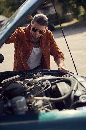 attractive young man in brown shirt with accessories looking attentively at engine of his carの写真素材