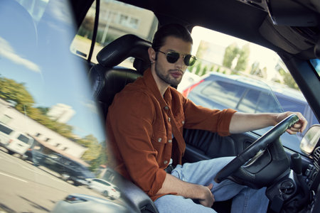 young good looking man with stylish sunglasses in vibrant attire posing in car and looking at camera