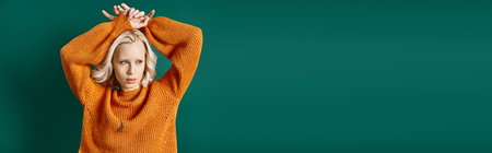 young blonde woman in mustard yellow sweater posing with hands above head on turquoise, bannerの写真素材