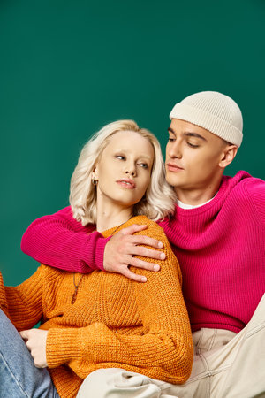 stylish couple in sweaters, man in beanie hugging blonde woman while sitting together on turquoiseの写真素材