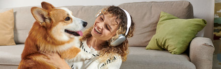 curly woman in wireless headphones playing with cute corgi dog in modern apartment, happy momentsの写真素材