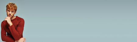 thoughtful man in red warm sweater with hand near chin looking at camera, fashion concept, bannerの写真素材