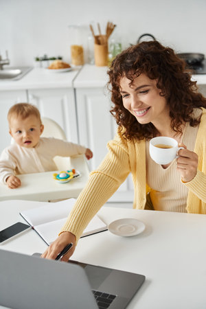 female freelancer with coffee cup working on laptop near toddler child in kitchen, multitaskingの写真素材