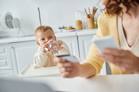 little girl drinking from baby bottle near mom with smartphone and credit card on blurred foreground