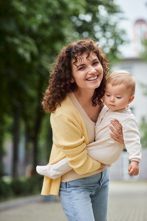 curly woman smiling at camera while holding little daughter on urban street, happy modern familyの写真素材