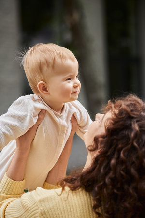 woman with wavy hair holding pleased little daughter in romper while spending time outdoors