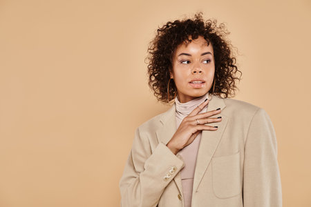 brunette african american woman with curly hair posing in turtleneck and blazer on beige backgroundの写真素材