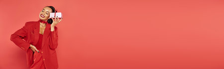 cheerful african american woman posing in suit with hand on hip and holding present on red, bannerの写真素材