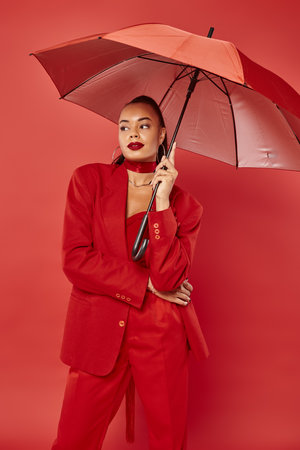 attractive young african american woman in jacket and pants standing under umbrella on red backdropの写真素材